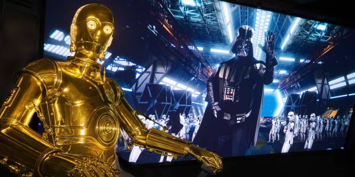 A gold humanoid robot stands in the foreground, while a large screen in the background displays a black-armored figure with a cape, surrounded by troops in white armor inside a space hangar, reminiscent of an epic scene you might encounter on a Disney World Vacation.
