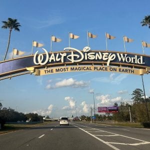 Entrance to Walt Disney World featuring a large archway with "The Most Magical Place on Earth" written below. Mickey and Minnie Mouse are displayed on pillars. Palm trees line the road under a blue sky.