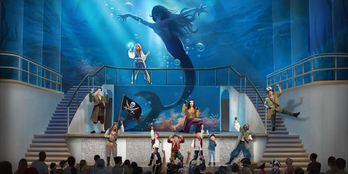 A theatrical performance features actors dressed as pirates on stage with a large background of a mermaid underwater. The set includes a pirate flag and marine-themed elements. An audience is visible at the bottom.
