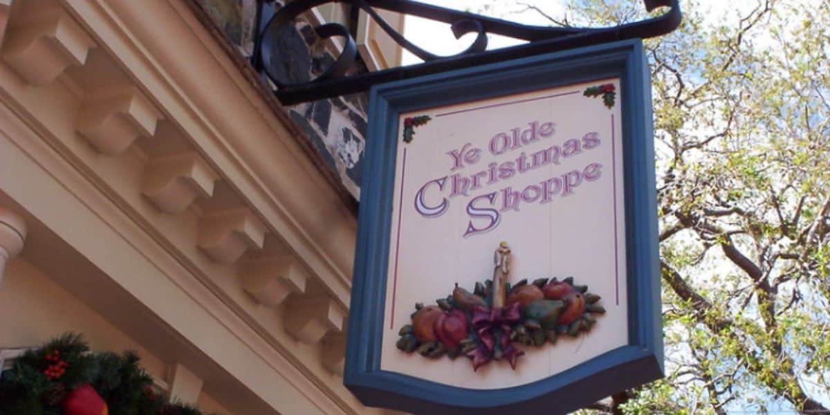 A storefront sign reads "Ye Olde Christmas Shoppe" in decorative lettering, adorned with festive holly and fruit illustrations. The sign hangs on a wrought iron bracket outside a building, with a clear sky and tree branches in the background.