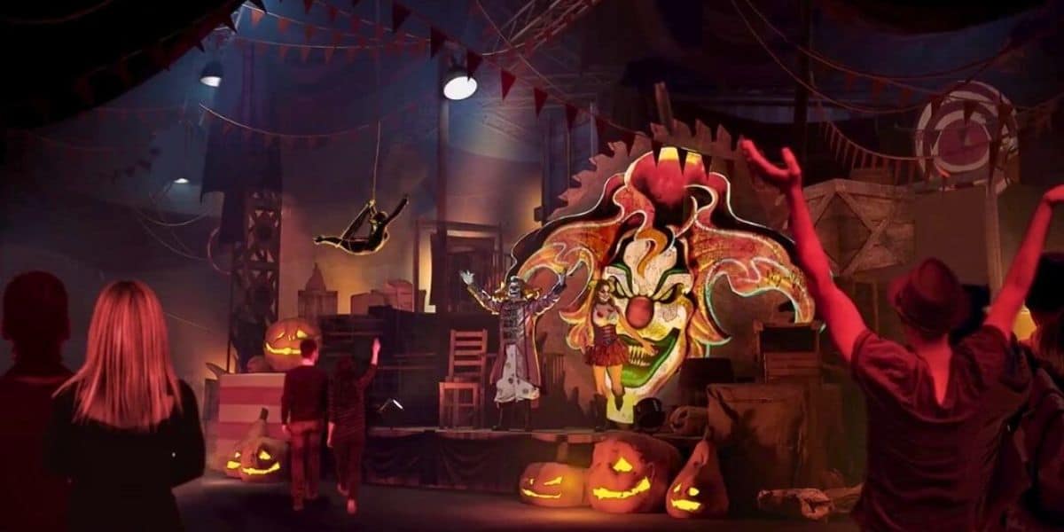 A dimly lit circus scene features a figure swinging on a trapeze, a large scary Jack the Clown image, and pumpkins with carved faces. People stand in the foreground, watching the performance as eerie lighting and decorations enhance the spooky atmosphere inside Universal Horror Unleashed.