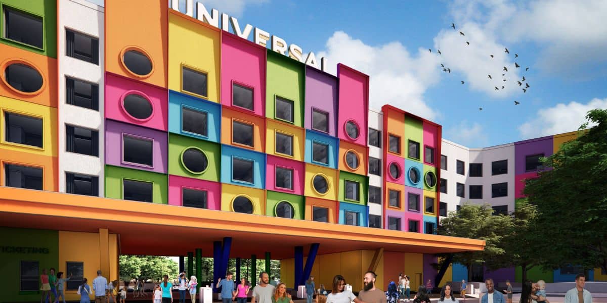 A vibrant, colorful building with a large "UNIVERSAL" sign resembles something you'd find at a Disney theme park. Multicolored geometric shapes decorate the facade. People gather at the entrance while birds soar in the clear blue sky above, with trees visible to the right.