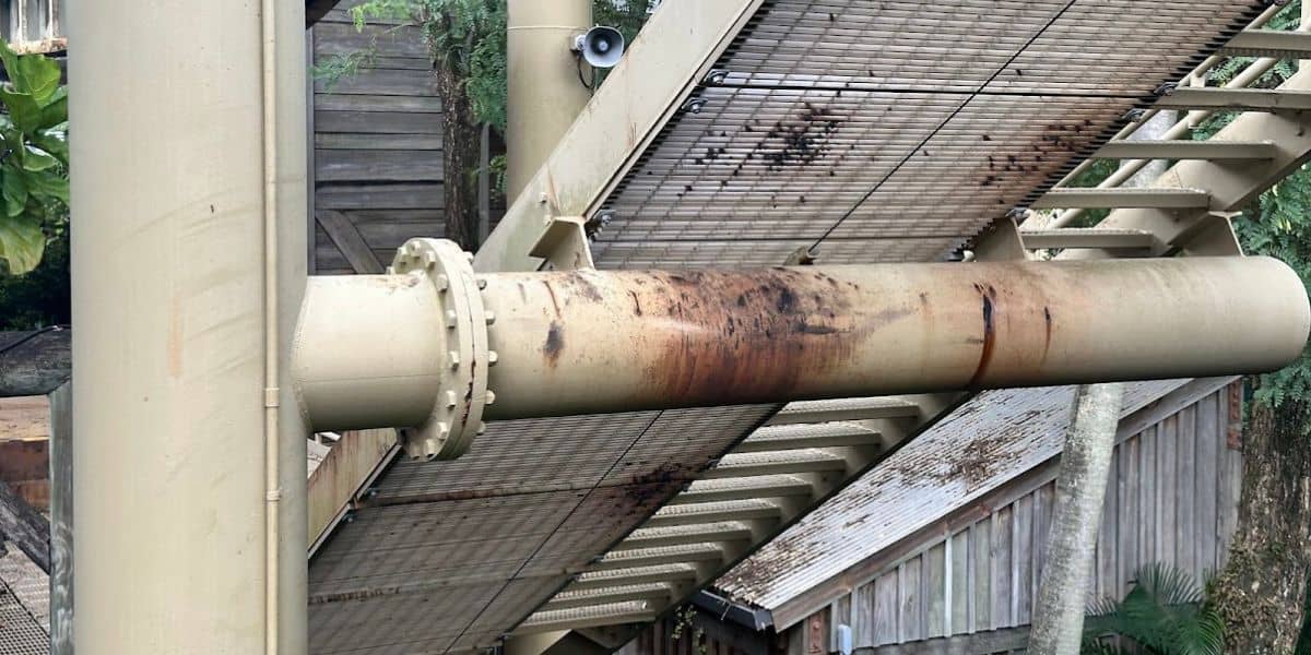 A rusty cylindrical metal pipe extends horizontally beneath a grated walkway, reminiscent of an opening day attraction. The structure is supported by beams and bolts, while in the background, trees stand alongside a wooden building crowned with a corrugated metal roof on Pteranodon Flyers at Universal Orlando Resort.