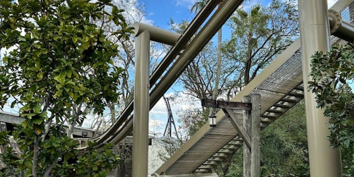 A roller coaster track with beige supports winds through a lush landscape of green trees. The sky above is blue with some clouds, creating a striking contrast with the foliage—a true universal attraction that resurrects thrill and wonder for all who ride.