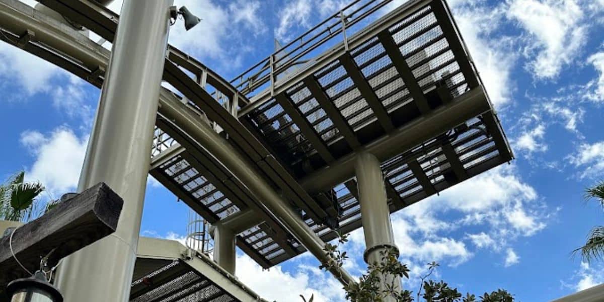 A low-angle view of the steel roller coaster track soars into a blue sky dotted with scattered clouds. This universal opening day attraction showcases its industrial beams and grated walkways, enveloped by lush green foliage at the base.