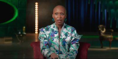 A person with a shaved head is sitting in a well-lit room, wearing a wickedly vibrant floral-patterned outfit. The background is softly blurred, showcasing vertical lights and a hint of furniture.