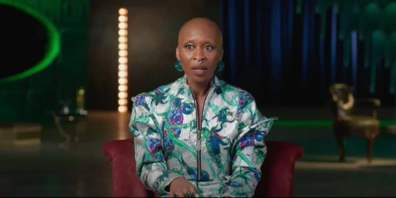 A person with a shaved head is sitting in a well-lit room, wearing a wickedly vibrant floral-patterned outfit. The background is softly blurred, showcasing vertical lights and a hint of furniture.