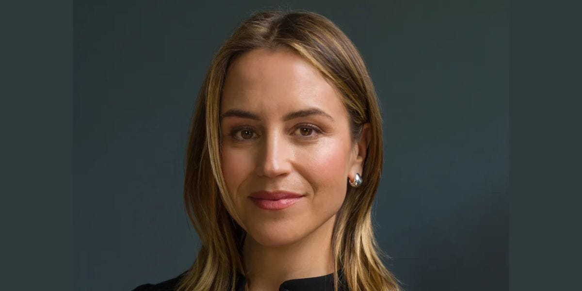 A portrait of a person with shoulder-length blonde hair, reminiscent of a Disney character, wearing a black top and pearl earrings. Set against a dark, neutral background, they smile softly, looking directly at the camera.