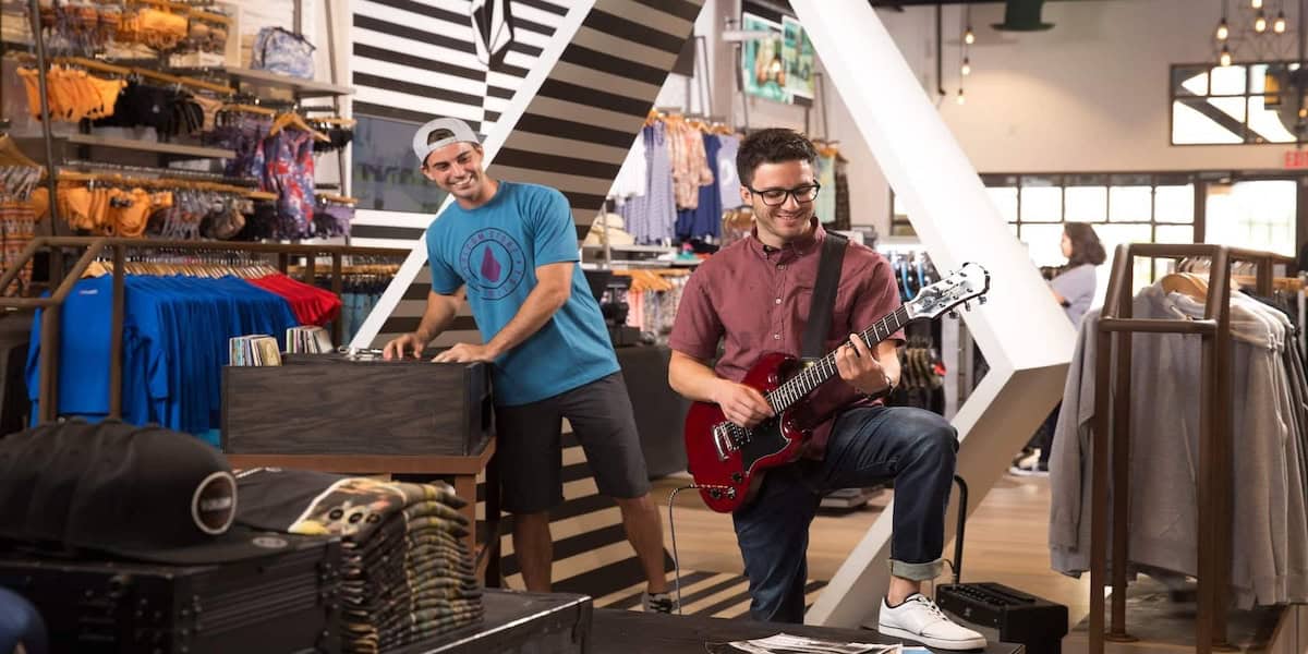 Two men inside a clothing store at a Disney World location, one playing a guitar while the other stands behind a DJ booth. Clothes are displayed in the background, creating a lively atmosphere.