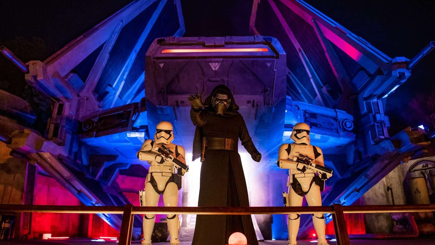 At Galaxy’s Edge, Kylo Ren stands in front of a Tie fighter, flanked by two First Order stormtroopers. The dramatic scene is enhanced by the red and blue lights casting an otherworldly glow.