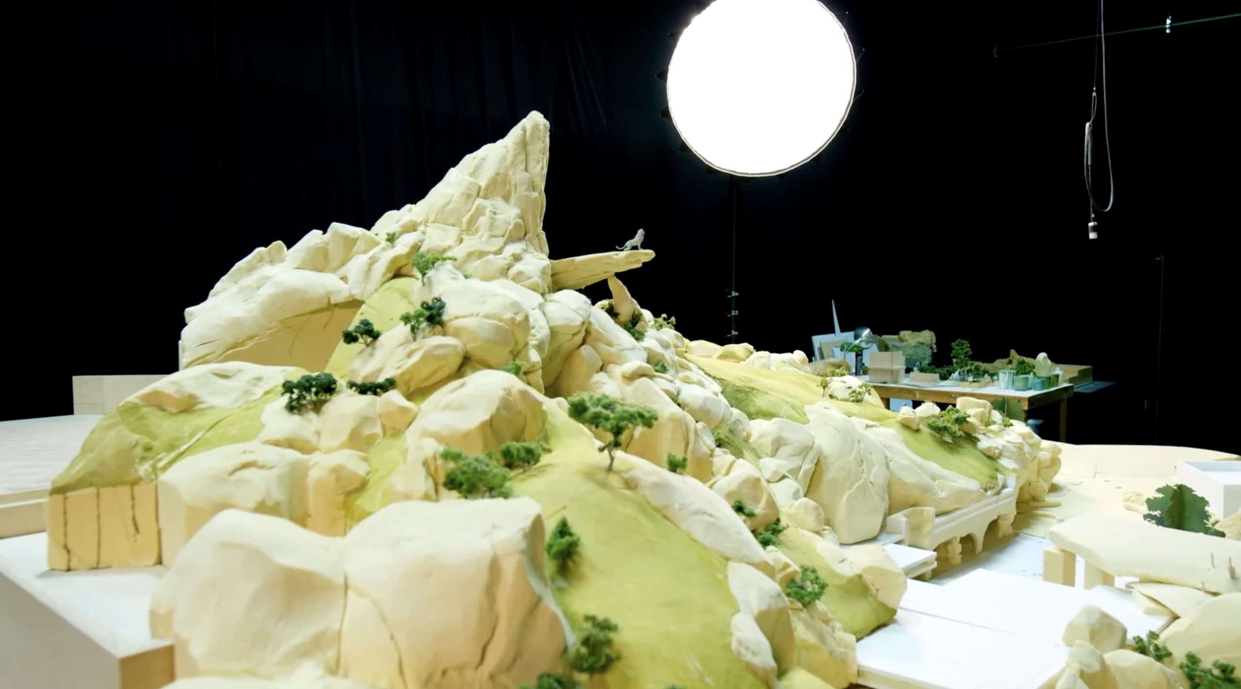 A detailed model of a mountainous landscape reminiscent of Splash Mountain with rocky formations and scattered green trees is set up under studio lighting. In the background, more miniature landscape elements and equipment evoke a thrilling attraction against a dark backdrop at this Disney Park in France, a new Lion King ride emerges.