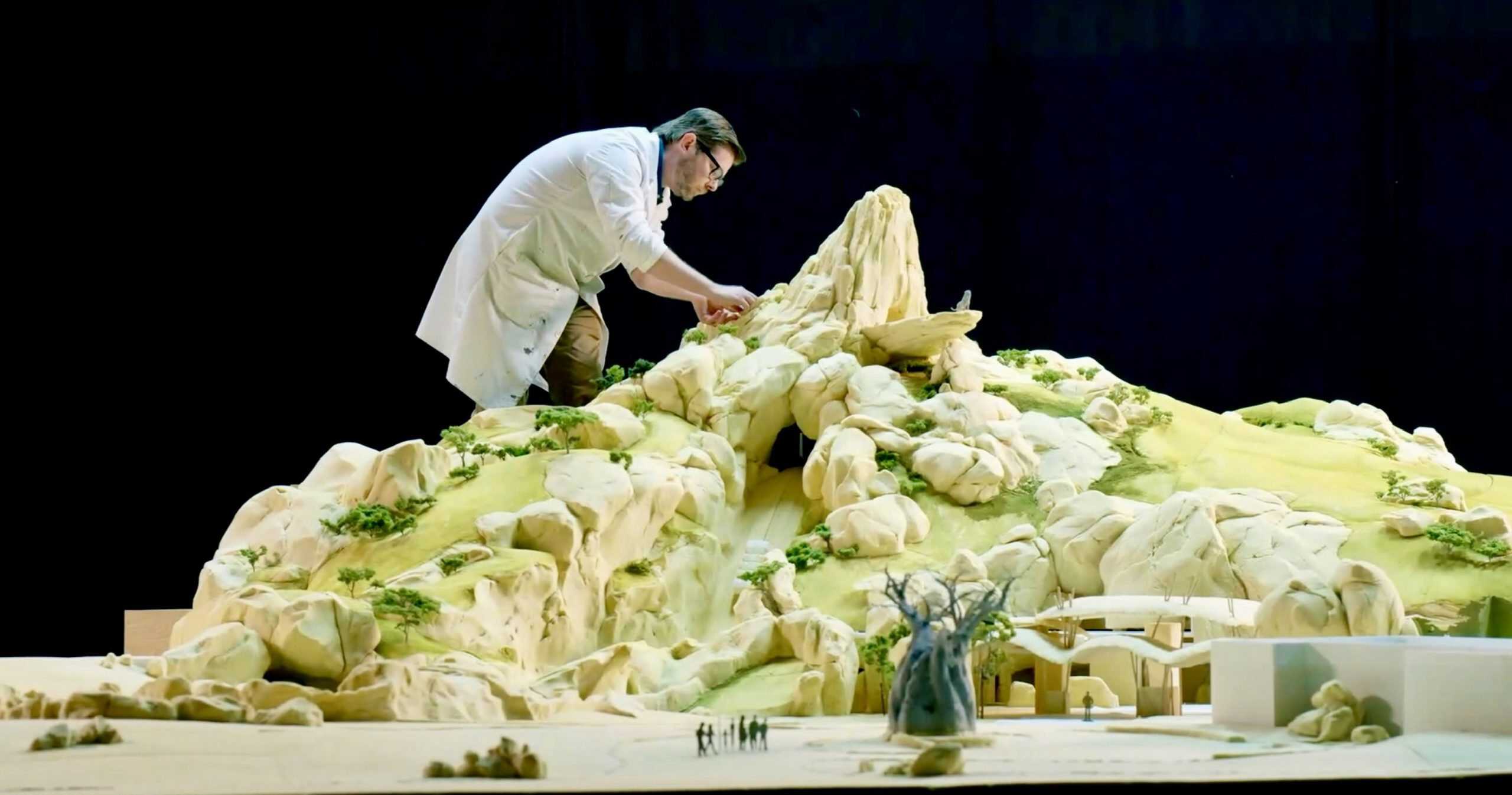 A person in a white lab coat is meticulously crafting a detailed model reminiscent of Splash Mountain, with rocky terrain. The studio's dramatic lighting highlights the intricate landscape and small structures at the base, evoking an attraction full of adventure and storytelling. Disney Park Lion King ride.