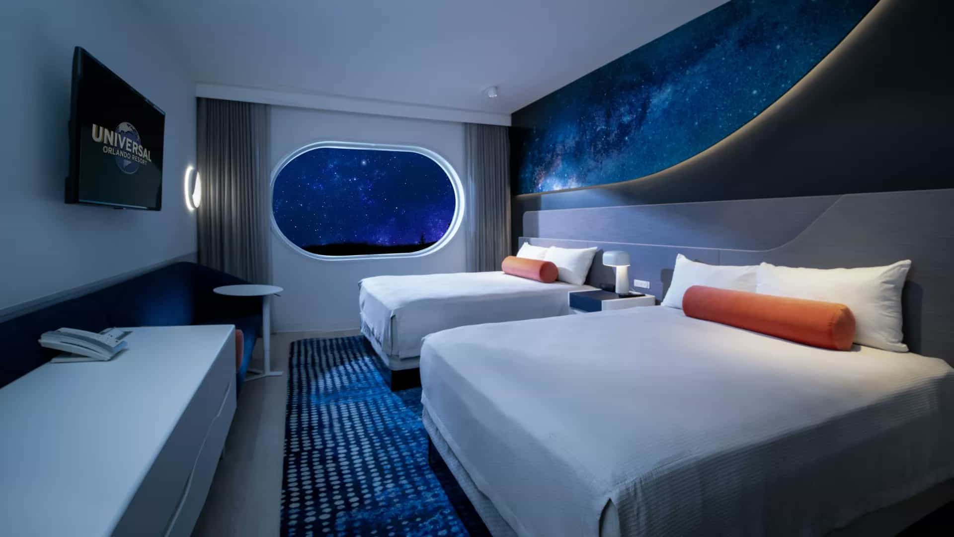 A modern hotel room features two double beds with white linens and orange cylindrical cushions. A wall displays a galaxy mural and a window shows a starry sky. A TV and a phone are on one side, and a blue patterned rug covers the floor.