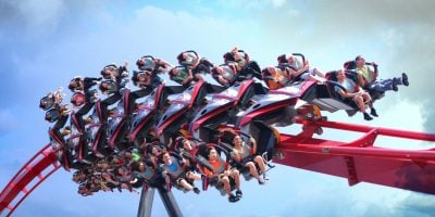 The X2 roller coaster with multiple rows of riders twists through the sky against a backdrop of clouds. The passengers show a mix of excitement and thrill, with their hands up or gripping the restraints. The coaster is red with black and silver accents at Six Flags Magic Mountain.