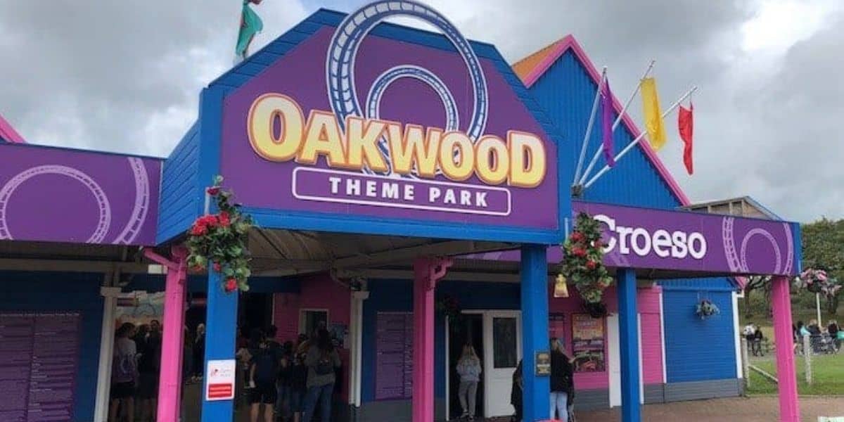 Entrance to Oakwood Theme Park, featuring a purple and blue facade with a large sign displaying the park's name. Roller coaster tracks are illustrated on the sign. People are gathered near the entrance. Flags are flying above.