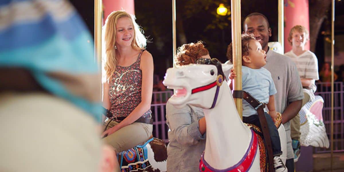 A group of people, including a young woman and a man holding a child, are smiling and riding on a carousel with white horses at the theme park. The festive background, filled with bright lights and blurred motion, adds to the enchanting experience.