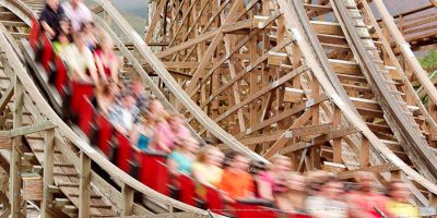 A group of people, blurred from motion, race through a theme park on a wooden roller coaster in red cars. The structure looms large as the riders, thrilled and excited, speed along the track.