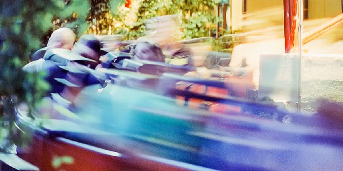 A blurred image captures the exhilarating speed of a theme park ride with passengers. The motion blur highlights excitement, while surrounding greenery and bright lighting enhance the vibrant atmosphere.