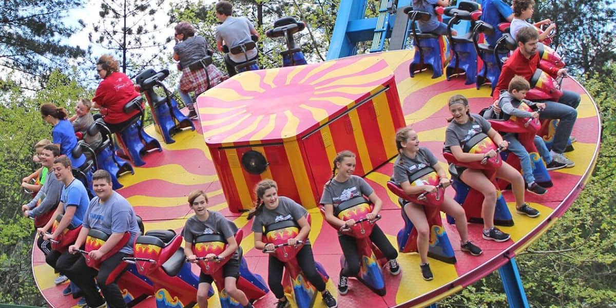 People are riding a colorful amusement park ride that spins in the air. The ride is circular with striped red and yellow patterns. Riders, including children and adults, are seated around the edges, some smiling while others hold on. Trees are in the background.