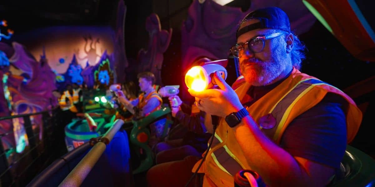 A person wearing glasses, a hat, and a reflective vest is holding a glowing toy blaster on a dark, colorful Disney World ride. Other guests are in the background, enjoying the vibrant and whimsical setting before its closure.