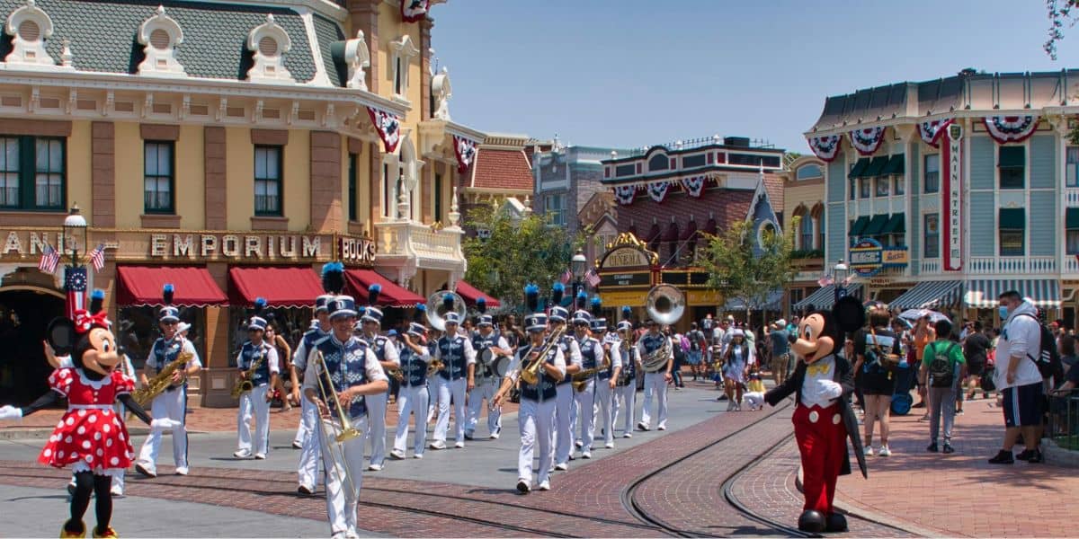 A marching band in blue uniforms parades down a street at the Disneyland Park. Mickey and Minnie Mouse, in their iconic outfits, wave to the guests. People, adhering to the dress code, watch from the sidewalks as buildings are adorned with patriotic bunting.