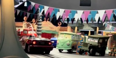 A lively scene depicts animated cars in a festive street setting. Brightly colored flags hang overhead. Various car models, including a van and a jeep, are visible, with whimsical buildings in the background.