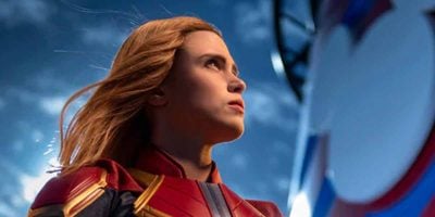 A woman in a red and gold superhero suit looks determinedly into the distance, with part of a round, blue, white, and red shield visible—celebrating Marvel Day at Sea on the Disney Cruise Line 2026—against a vibrant blue sky.