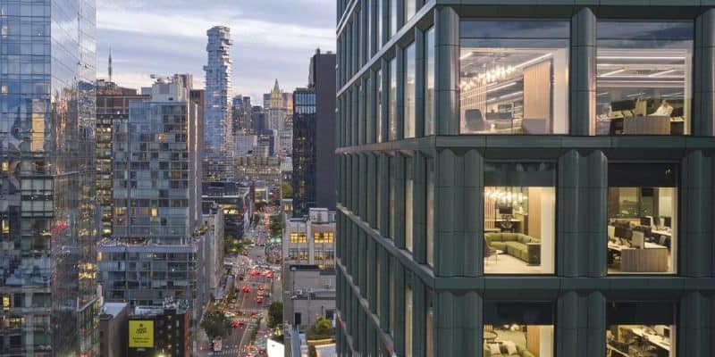 A cityscape at dusk features modern high-rises, including the iconic Disney headquarters. The focus is on a glass skyscraper with illuminated interiors showing office and residential spaces. The street below bustles with traffic and lights, while more urban architecture looms in the background.