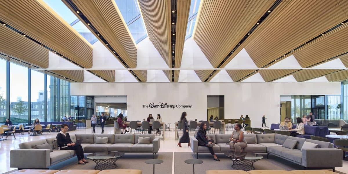 The modern lobby at Disney headquarters boasts high ceilings and wooden panels, with "The Walt Disney Company" logo prominently displayed. People converse on gray couches while others gather near tables. Large windows flood the space with natural light, creating an open, airy ambiance.