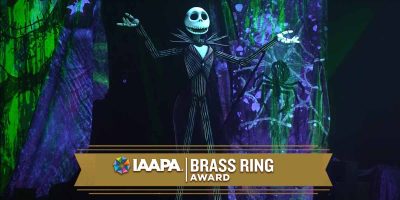 Jack Skellington, a tall skeleton in a pinstripe suit, stands on stage with arms raised. The background is decorated with green and purple spooky designs. A banner reads “IAAPA Brass Ring Award.”.