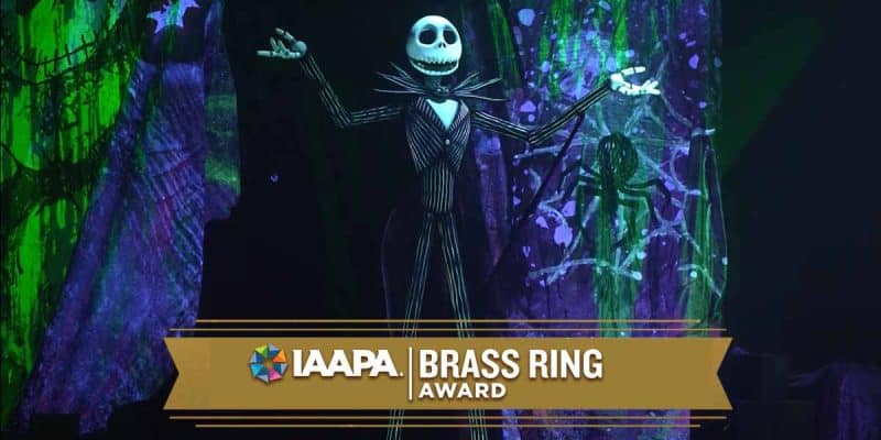 Jack Skellington, a tall skeleton in a pinstripe suit, stands on stage with arms raised. The background is decorated with green and purple spooky designs. A banner reads “IAAPA Brass Ring Award.”.