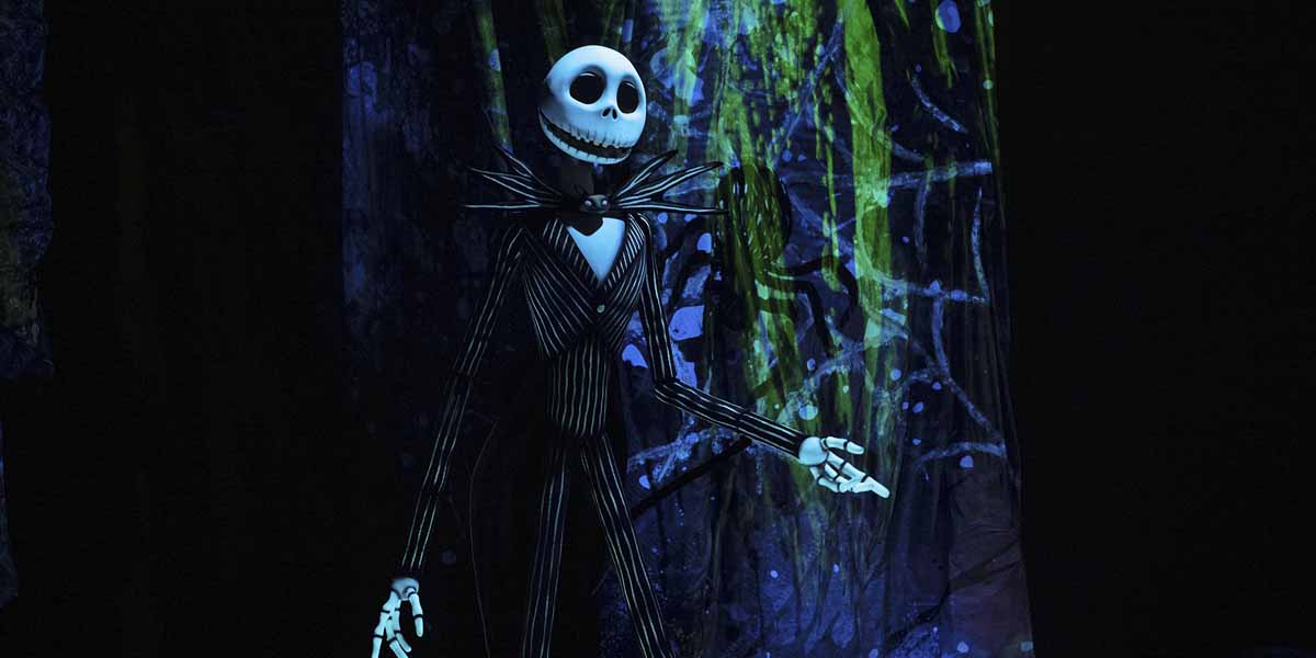 A tall skeleton figure in a black and white pinstripe suit stands with outstretched hands against a dark, abstract, and colorful background. The character has a large, round skull and a wide, smiling mouth.