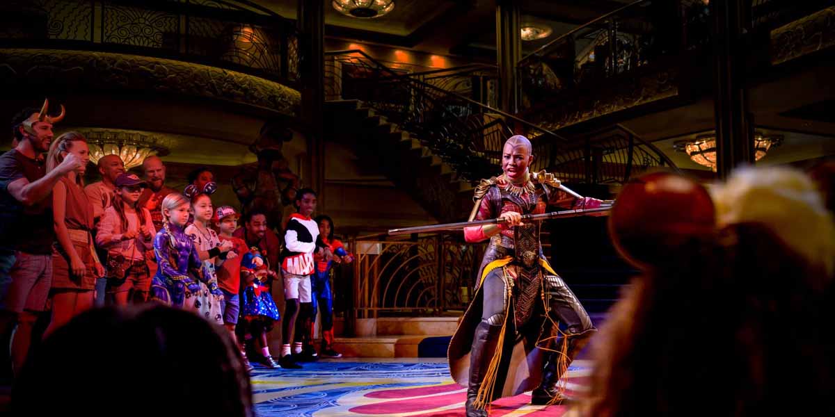 A performer in an elaborate warrior costume demonstrates with a spear before children and adults on a grand staircase, under warm lighting—an exciting moment during Marvel Day at Sea aboard Disney Cruise Line 2026.