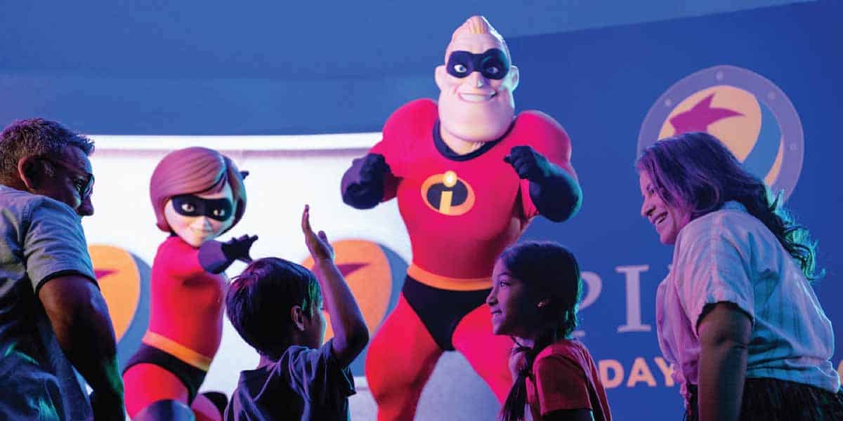 Two adults and two children smile and interact with Mr. Incredible and Elastigirl at a Pixar Day at Sea event, surrounded by Pixar-themed decor on the Disney Cruise Line 2026.