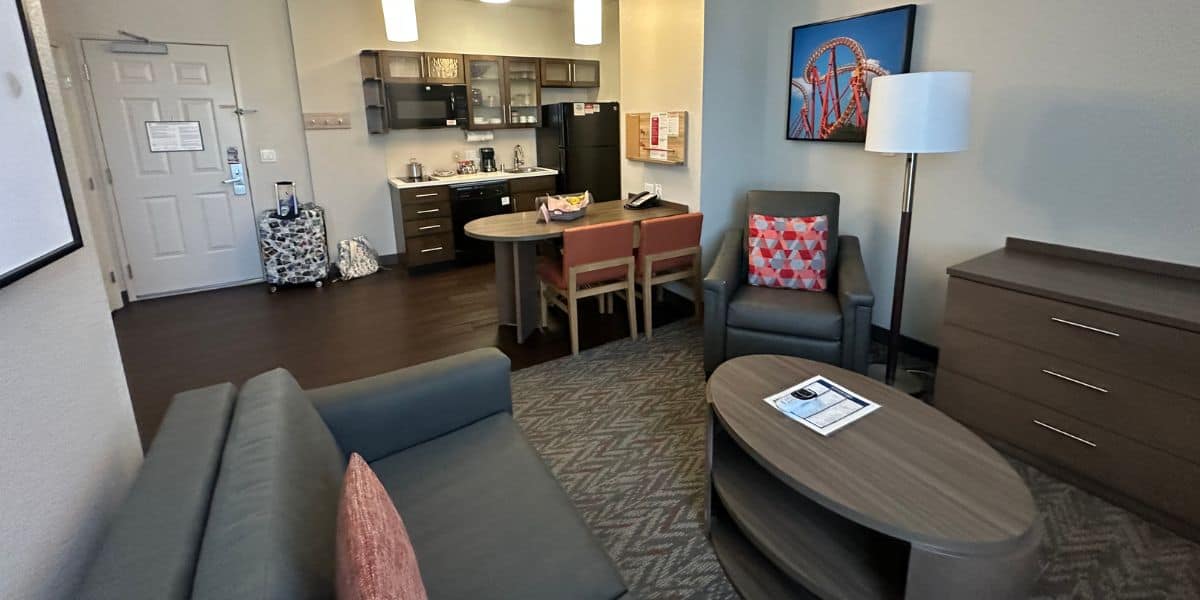 A cozy Disneyland Hotel suite with a small kitchen, dining table for two, armchair, sofa, coffee table, dresser, and luggage near the door. The modern decor is neutral with colorful accents for a welcoming stay.