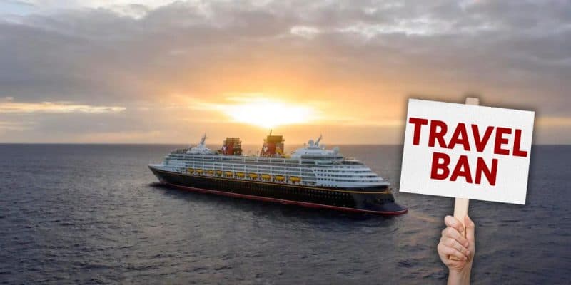 A large cruise ship sails on the ocean at sunset while a hand in the foreground holds a sign that reads "TRAVEL BAN" in bold red letters, which i coming to select locations for the Disney Cruise Line.