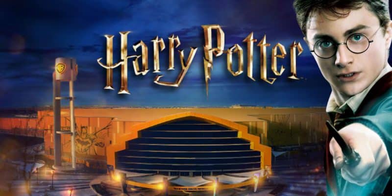 The Harry Potter logo above a large, modern building at night with a clock tower and the Warner Bros. logo; a young man with glasses and a wand points forward on the right inside of the WB Dubai theme park in the Middle East.