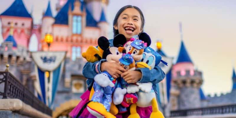 A joyful child holds several Disney character plush toys in front of a fairy-tale castle at Disneyland Resort. The background features turrets and decorations, enhancing the celebration atmosphere. The child is smiling widely, wearing a denim jacket.