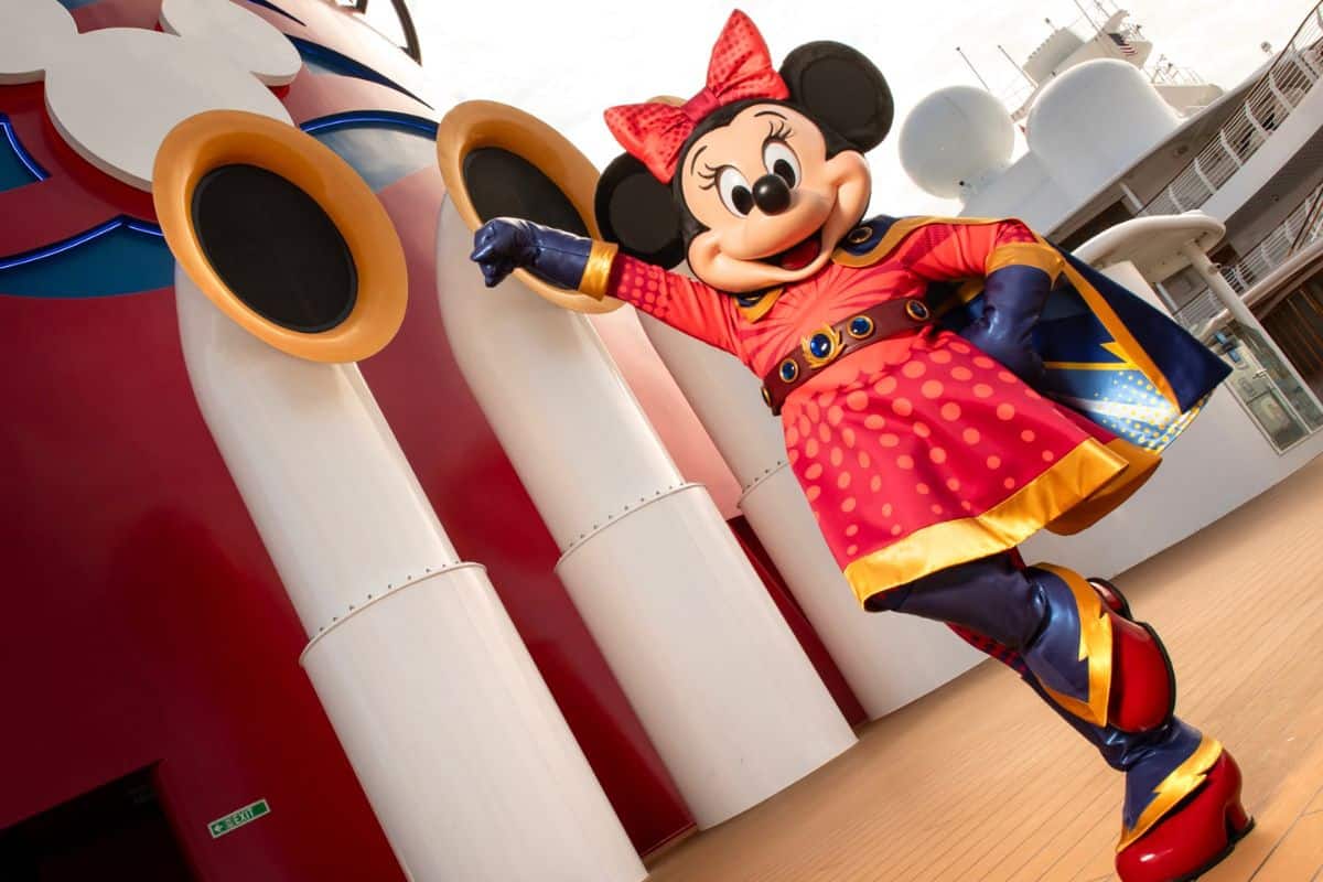 Minnie Mouse, in a vibrant red superhero costume with a golden cape, energetically poses on the deck of the Disney Destiny.