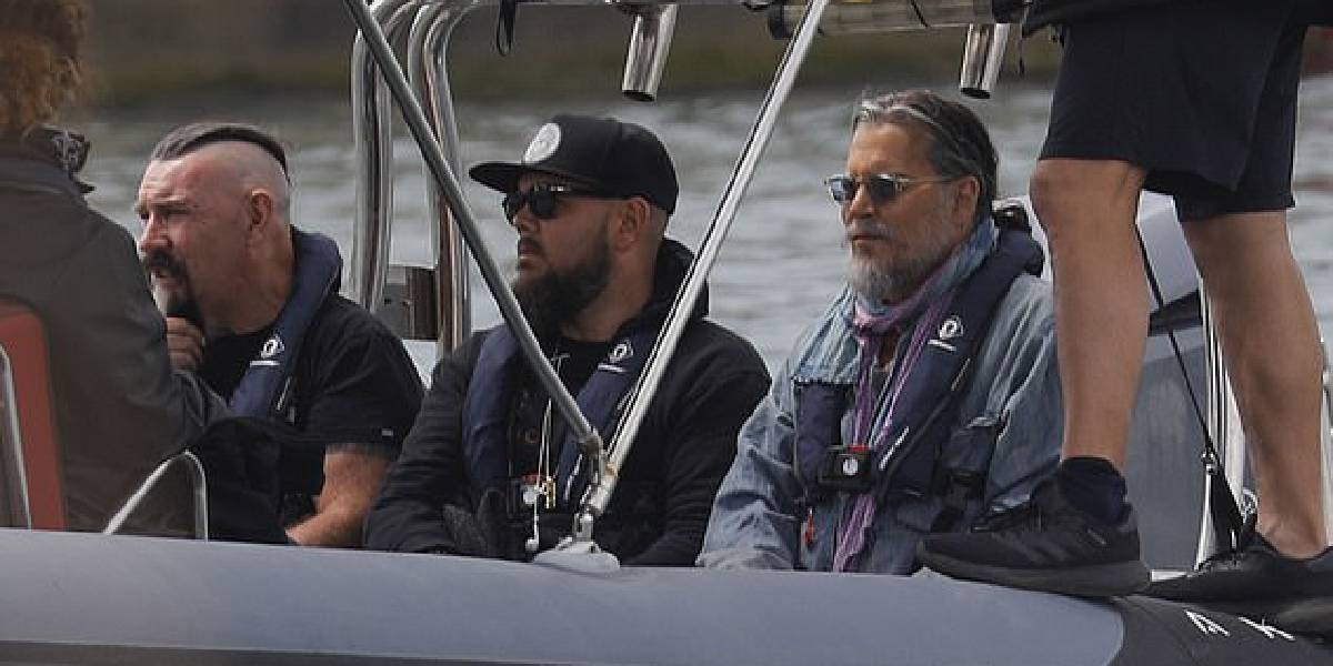 Three men in life jackets sit on a boat, resembling a Pirates franchise scene. One sports a mohawk, another sunglasses and a cap, while the third has gray hair and a beard. Another person's legs in shorts and sneakers hint at possible filming with Johnny Depp vibes.