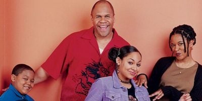 Four people stand smiling in front of a peach-colored wall, posing cheerfully like a Disney Channel cast. A young boy leans left, with a tall man in red behind him; a teen girl and woman with braids stand close by.