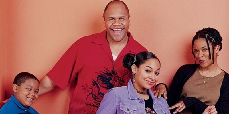 Four people stand smiling in front of a peach-colored wall, posing cheerfully like a Disney Channel cast. A young boy leans left, with a tall man in red behind him; a teen girl and woman with braids stand close by.
