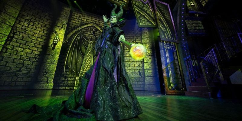 A performer dressed as Maleficent stands on stage in a dramatic pose, holding a glowing orb, surrounded by green and dark lighting and gothic castle scenery during Disney Villains: Unfairly Ever After.
