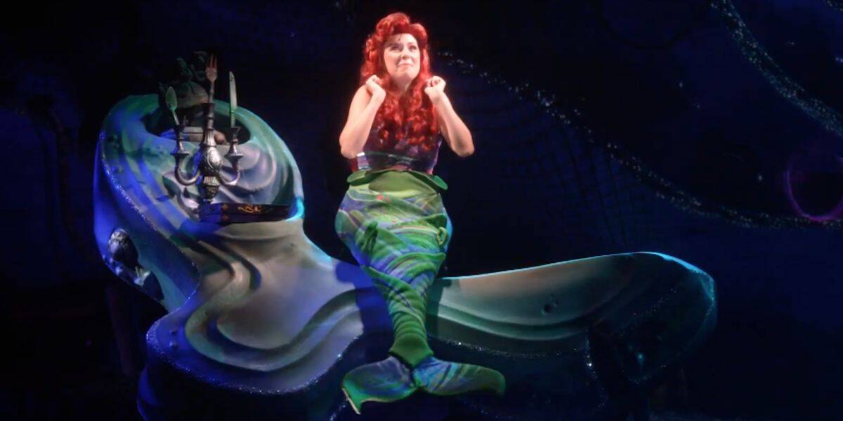 A performer dressed as Ariel from "The Little Mermaid" musical at Walt Disney World sits on a large seashell prop, wearing a red wig and green mermaid tail costume, smiling brightly under stage lights.