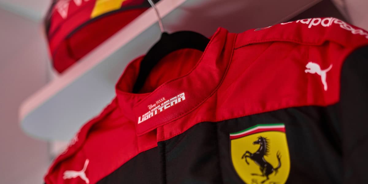 A close-up of a red and black Ferrari racing suit, featuring the Ferrari logo with a prancing horse, Puma logo, and "LECLERC" text. A matching Ferrari cap sits on a shelf in the background—perfect for the Formula 1 enthusiast.