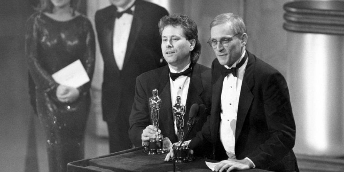 Two men in tuxedos stand at a podium holding Oscars, giving a speech at an awards ceremony. A woman and another man, also dressed formally, stand in the background.