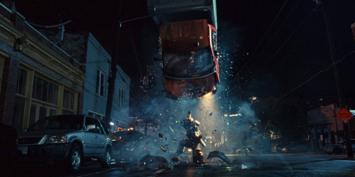 A Marvel superhero series icon stands in the middle of a dark city street at night, lifting a flaming car overhead as debris and sparks fly around, while parked cars and old buildings line the street—just like Ironheart in action.