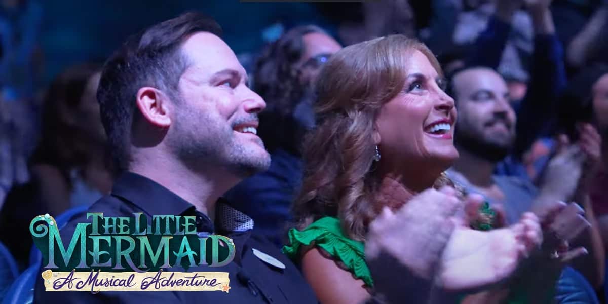 An excited audience claps and smiles at a live event in Hollywood Studios. In the foreground, a man in a dark jacket and a woman in a green dress enjoy the show. "The Little Mermaid: A Musical Adventure" logo is in the bottom left corner.