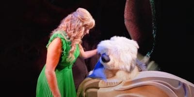 Under stage lights, a woman in a bright green dress gently pats a large, fluffy white and gray dog sitting in a wooden boat—reminiscent of Jodi Benson’s performances at Hollywood Studios in The Little Mermaid.