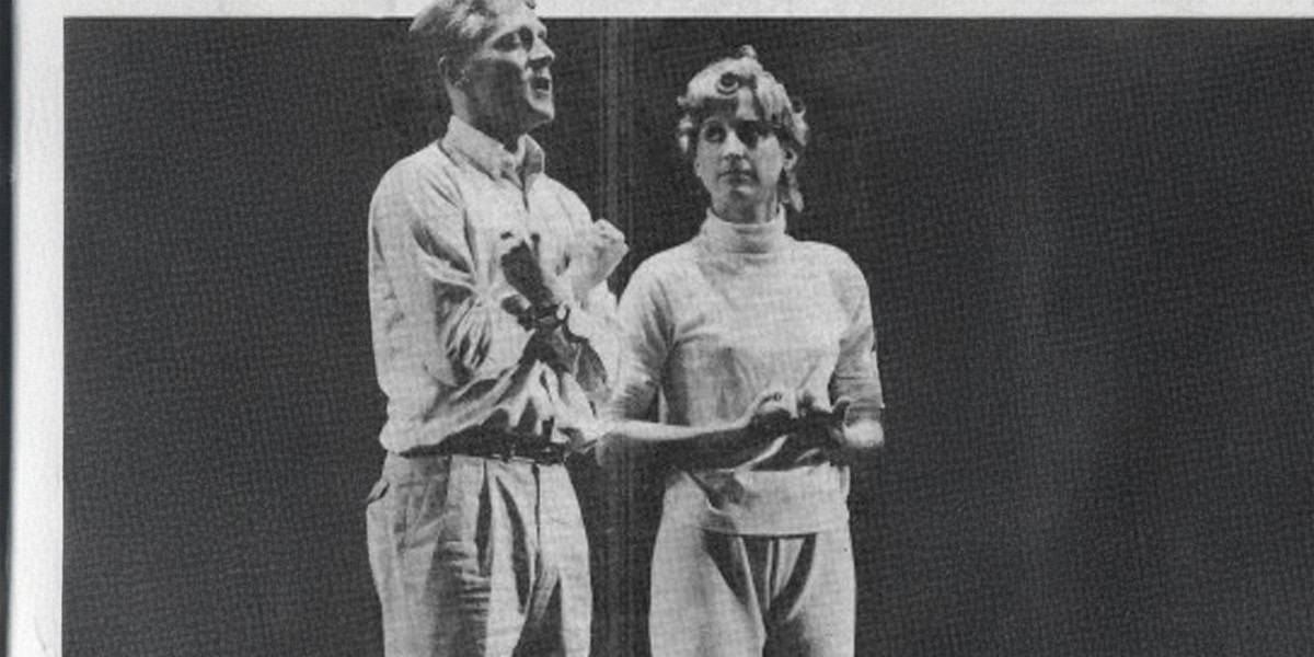 A black and white photo of Jodi Benson and Howard Ashman close together on stage, both with expressive hand gestures, appearing to perform or speak to an audience. The background is plain and dark.
